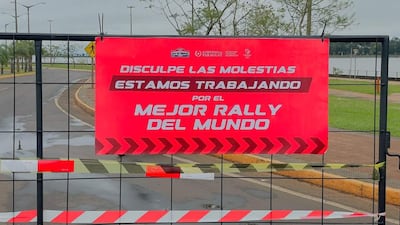 Cartel de advertencia en la vía pública con texto sobre el Rally de Paraguay, entorno urbano y áreas verdes visibles.