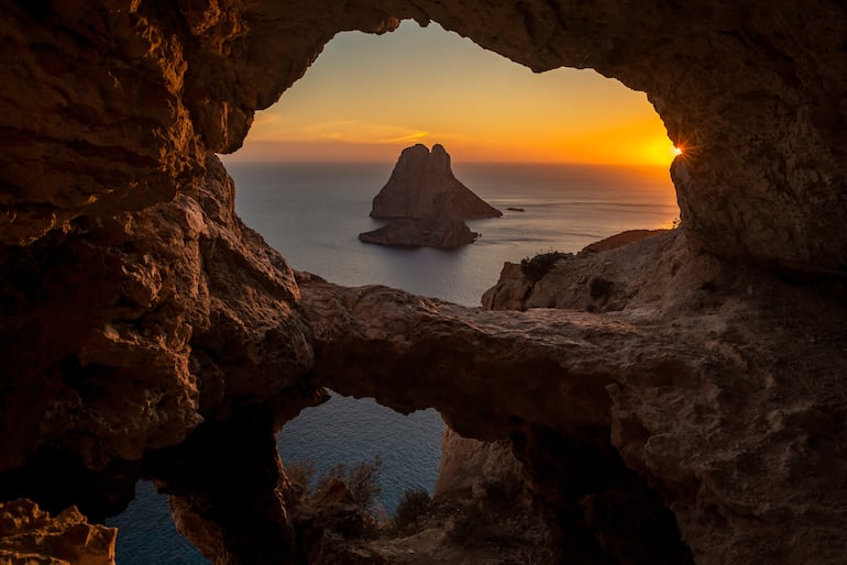 Vista del islote de Es Vedra a través de los agujeros rocosos de una cueva al atardecer, Sant Josep de Sa Talaia, Ibiza, Islas Baleares, España.