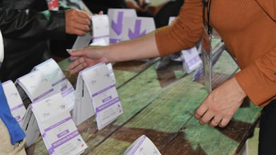 Manos de personas interactuando con boletas de votación organizadas en mesas de madera, en ambiente de votación interior.