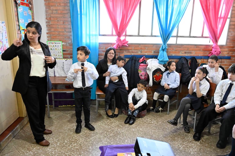 Mujer de pie gesticulando en aula, niño con corbata negra y varios niños atentos en el fondo, todos en uniforme escolar.