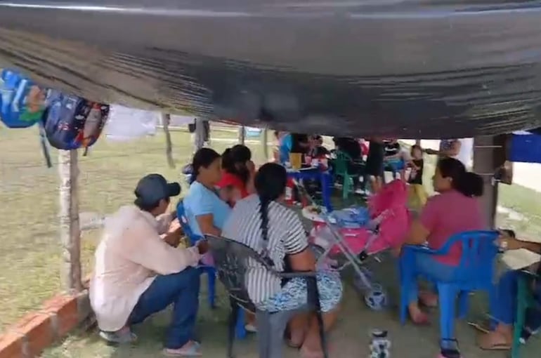 Grupo de personas sentadas bajo un toldo, algunas conversan, una mujer de espaldas con trenzas, carrito de bebé a la derecha.