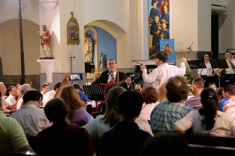 Hombre con gafas y guitarra al frente, mientras otros músicos siguen su dirección en un ambiente de iglesia decorado.