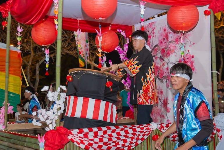 Cuatro jóvenes en atuendos coloridos, uno golpeando un tambor, rodeados de faroles rojos en un evento festivo.