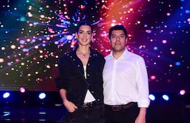 Mujer con conjunto de denim negro y camiseta blanca junto a hombre con camisa blanca, sonrientes en un ambiente festivo iluminado.