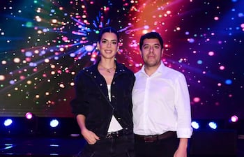 Mujer con conjunto de denim negro y camiseta blanca junto a hombre con camisa blanca, sonrientes en un ambiente festivo iluminado.
