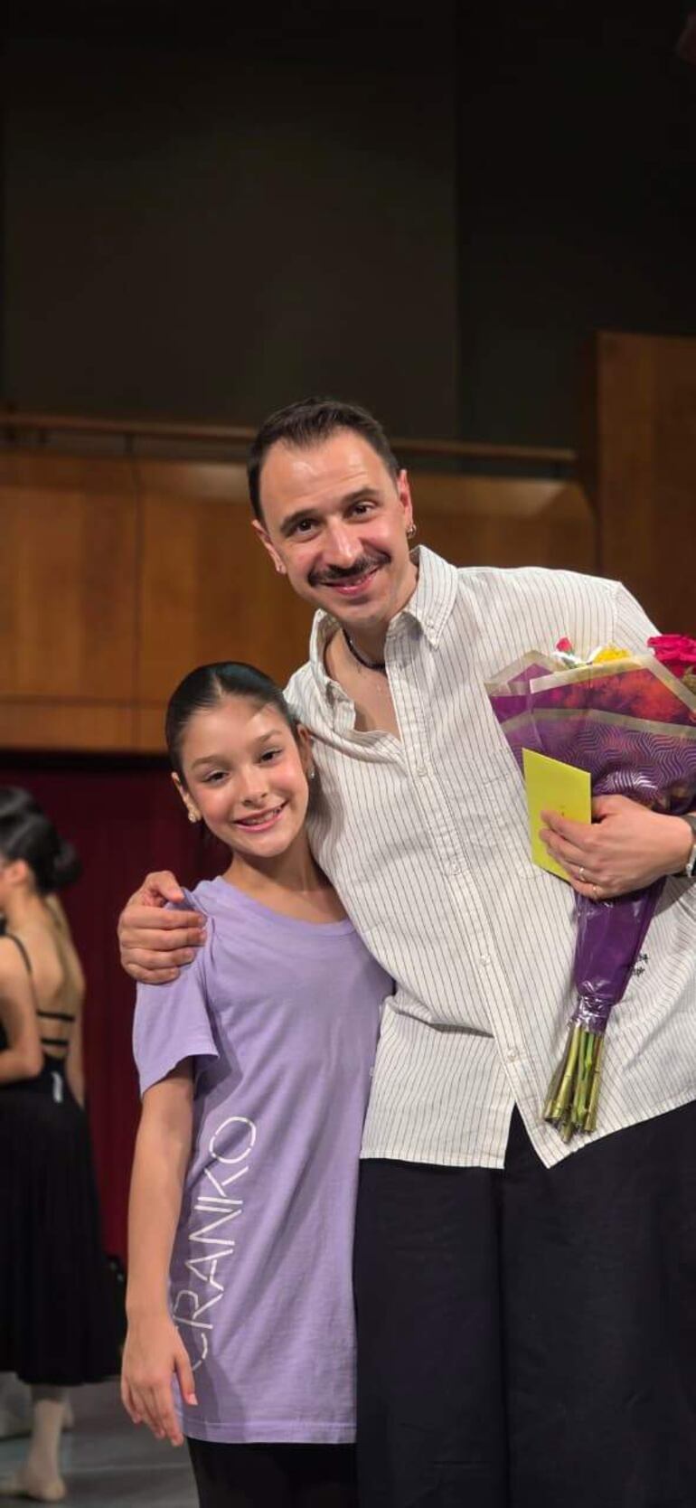 Niña sonriente en camiseta morada y hombre en camisa blanca sosteniendo un ramo de flores, rodeados de figuras en danza.