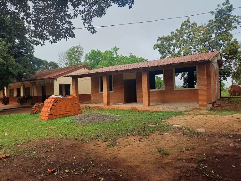Escena de construcción en el exterior de escuela rural con ladrillos y terreno expuesto rodeado de vegetación.