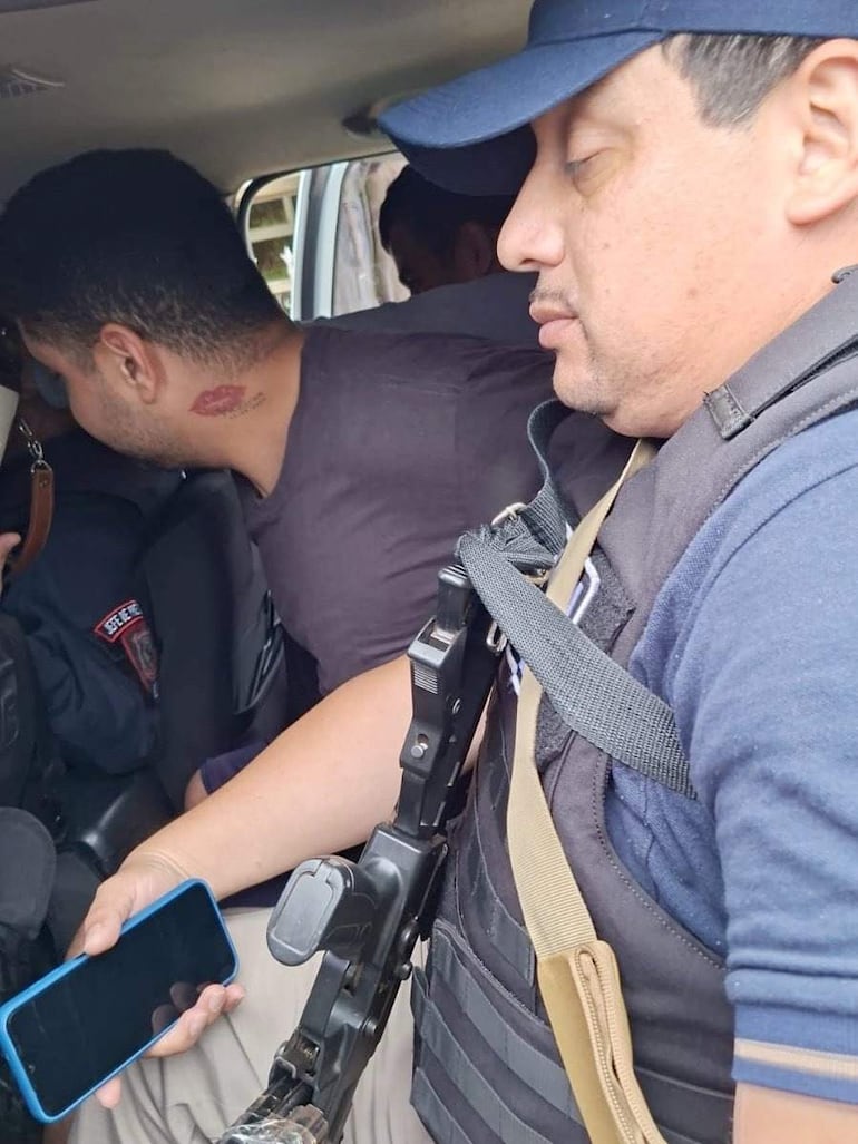 Hombre con camiseta negra y un tatuaje en el cuello junto a otro con gorra azul y chaleco táctico, sosteniendo un arma y un teléfono.