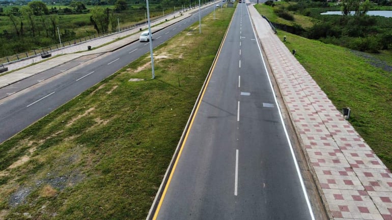 Vista aérea de la carretera con vehículos a la izquierda y acera pavimentada a la derecha, rodeada de áreas verdes.