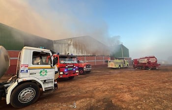 Camiones de bomberos en acción, rodeados de humo, atendiendo un incendio en un depósito de yerba canchada.