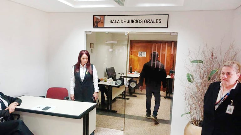 Mujer de pie con chaleco negro y camisa blanca en sala de juicios, otra mujer a su derecha con portafolio, hombre saliendo.