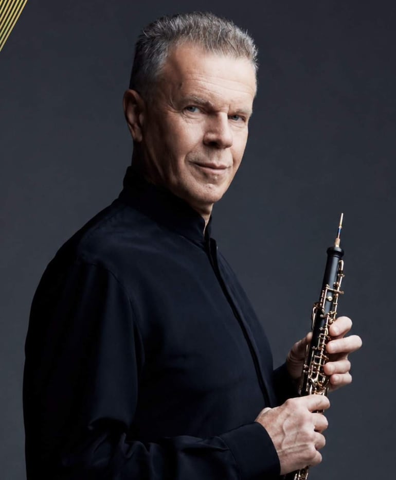 Alexei Utkin, hombre de mediana edad con cabello canoso y camisa negra, sostiene un oboe d'amore en un fondo oscuro.