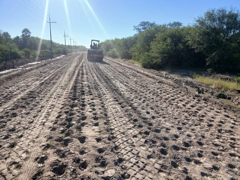 Camino de tierra con huellas y máquina pesada levantando polvo en un entorno rural con vegetación.