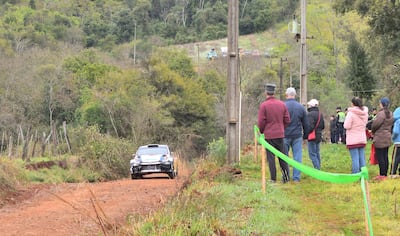 Espectadores con ropa abrigada observan un auto de rally en un camino de tierra, rodeados de vegetación.
