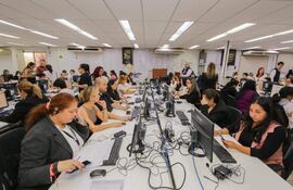 Una mujer con auriculares toma notas en una oficina iluminada, rodeada de otros empleados concentrados en sus tareas diarias.