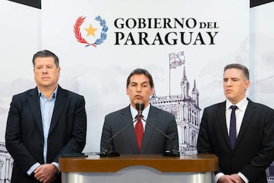 Javier Giménez en traje oscuro y corbata roja, flanqueado por Gustavo Villate y Enrique Biedermann, en conferencia de prensa formal.