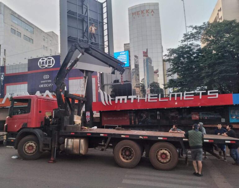 Seis personas observan un camión rojo con grúa que sostiene un cartel en un entorno comercial.