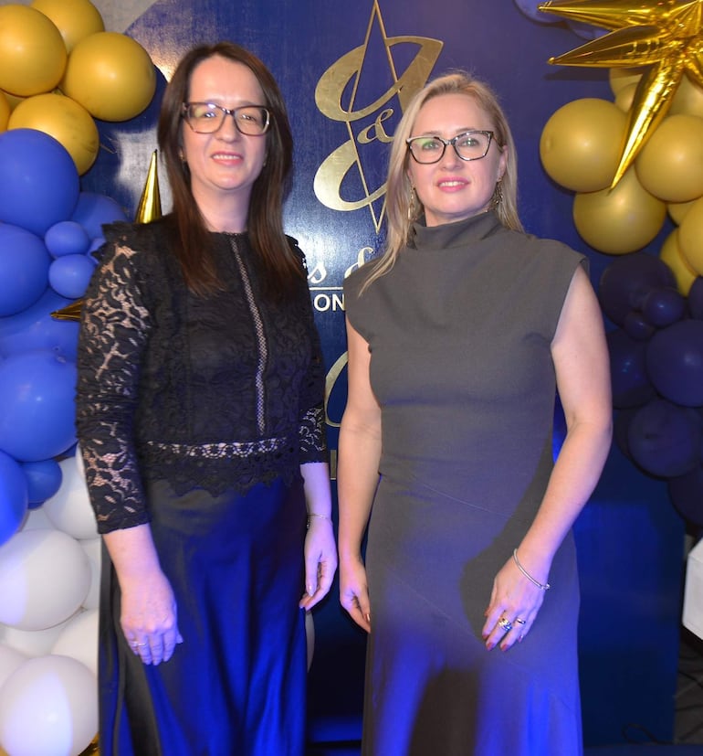 Mujeres luciendo vestidos elegantes, una en negro y otra en gris, en un ambiente decorado con globos y detalles dorados.