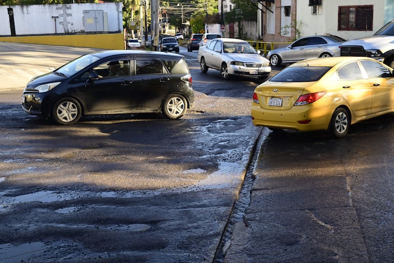 Intersección de calles urbanas con vehículos visibles y pavimento dañado, reflejando el impacto de la lluvia reciente.