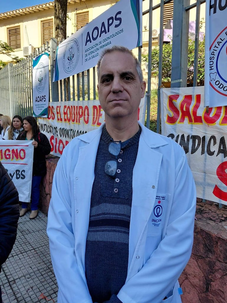 Gabriel Taddey con bata blanca, mirando a la cámara, mientras fondo sostiene carteles de protesta por la salud.