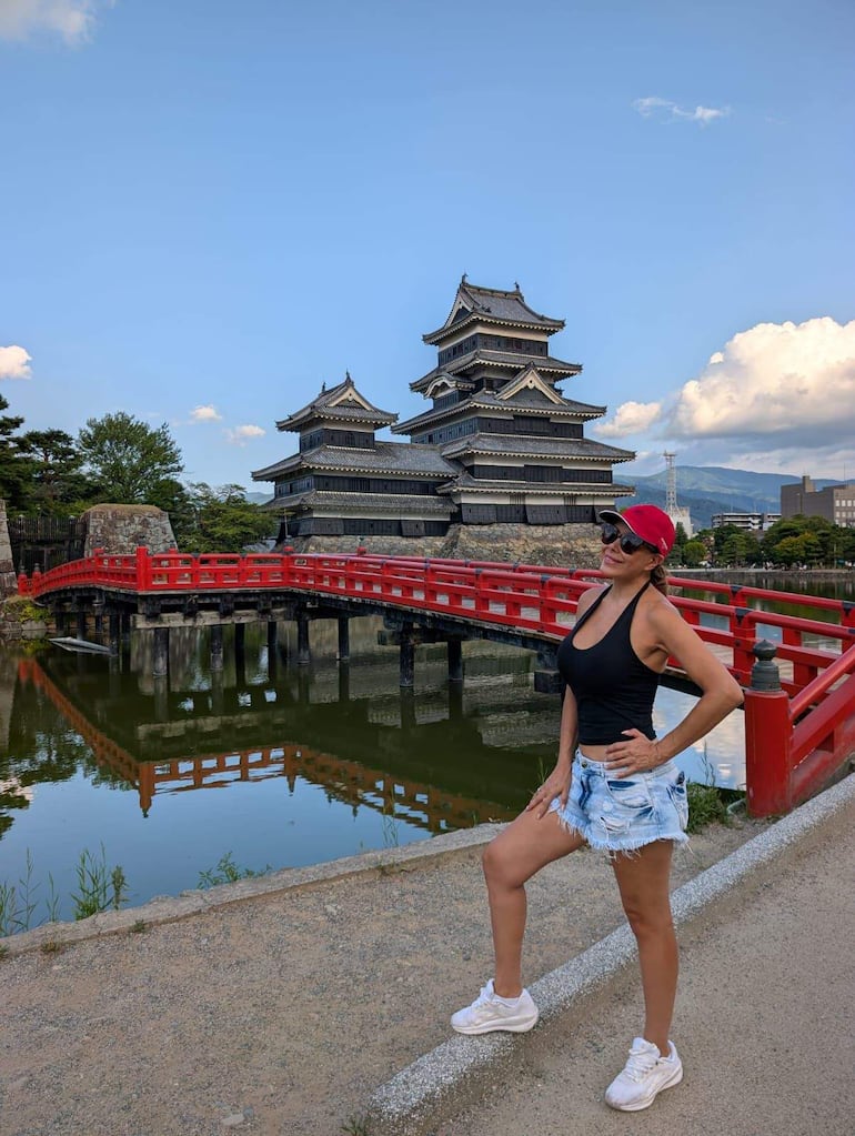 Mujer de cabello recogido con camiseta negra y short de mezclilla, sonriendo en un entorno natural frente al castillo Matsumoto-jō.