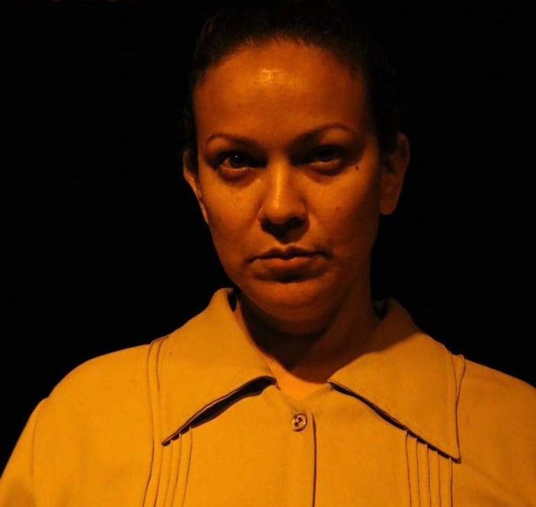 Elenco en escena, con vestimenta teatral y expresiones serias, ambientados en un escenario iluminado.