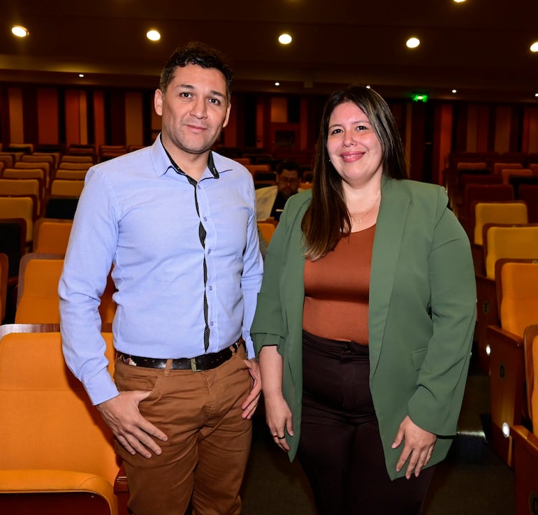 Hombre y mujer sonrientes en auditorio con asientos naranjas y marrones, ambos de pie en el primer plano.