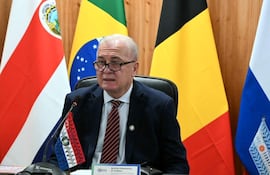 Dr. Emiliano Rolón Fernández en conferencia con camisa blanca, saco oscuro y corbata de rayas, junto a banderas de varios países.