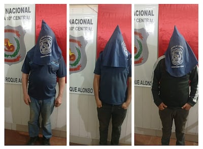 Tres hombres con capuchas azules de pie frente a una pared con el escudo de Mariano Roque Alonso en un ambiente formal.