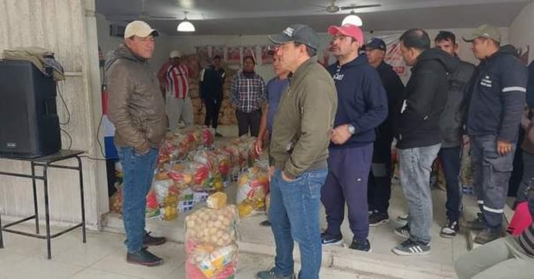 Hombre con chaqueta oscura y gorra lidera reunión, mientras otros lo escuchan atentamente entre bolsas de alimentos.