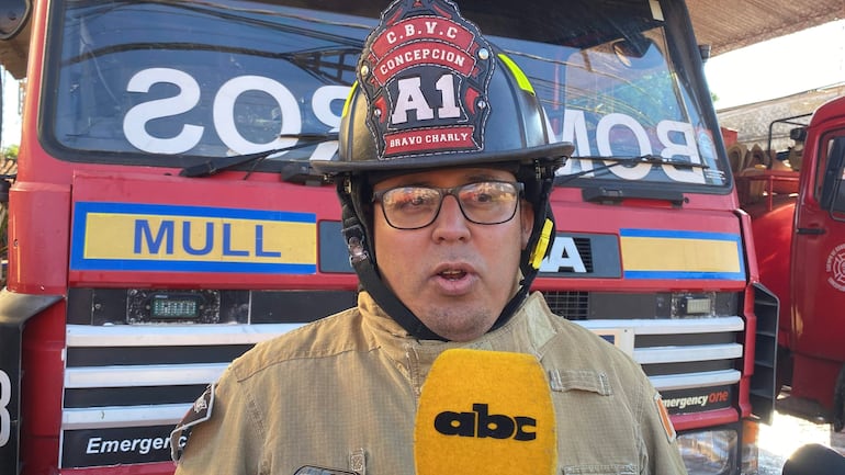 Bombero con casco y uniforme azul sostiene micrófono, frente a camión de bomberos rojo y amarillo, en un entorno exterior.