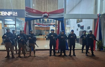 Ocho policías uniformados con armas largas, en guardia frente al edificio BNF 24 HORAS, con la bandera paraguaya en el entorno.