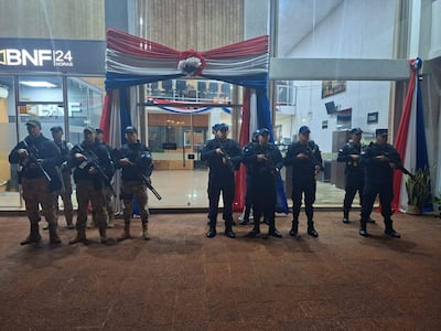 Ocho policías uniformados con armas largas, en guardia frente al edificio BNF 24 HORAS, con la bandera paraguaya en el entorno.