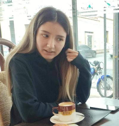 Joven con cabello largo y lacio en café, viste jersey oscuro, se toca el cabello y tiene taza de café en la mesa.