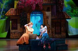 Mujeres en el escenario: una con vestido amarillo y otra con vestuario vibrante, en diálogo ameno entre decoraciones teatrales.