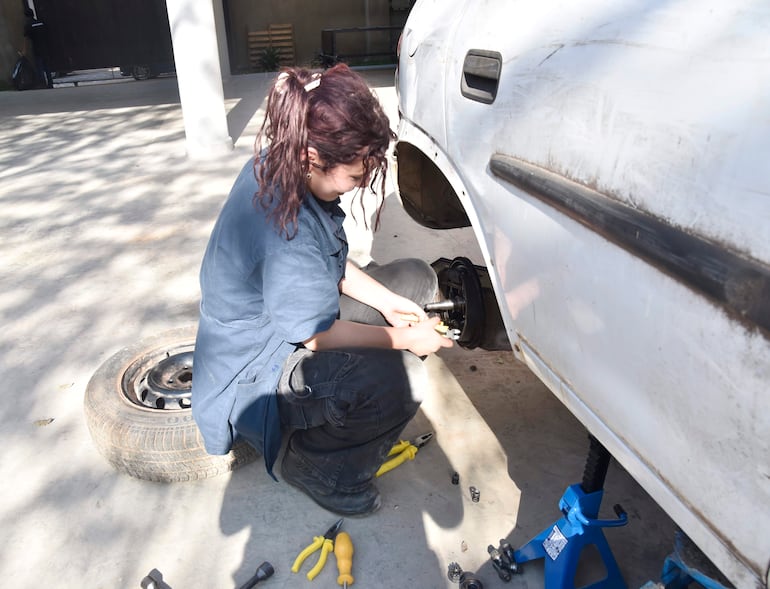 Mujer joven con camisa oscura y jeans, sentada sobre un neumático, usando herramientas en la mecánica de un auto blanco.