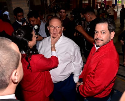 Raúl Latorre con gafas, camisa blanca y pantalón azul, interactúa con una mujer de cabello largo y vestido rojo en un ambiente iluminado.