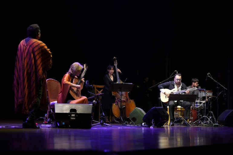 Berta Rojas con túnica naranja toca la guitarra, acompañada de cuatro músicos en un escenario con luz tenue.