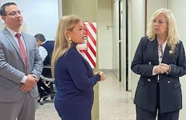 Mujeres en trajes formales discuten en ambiente de oficina. Una lleva abrigo azul oscuro y la otra traje negro.