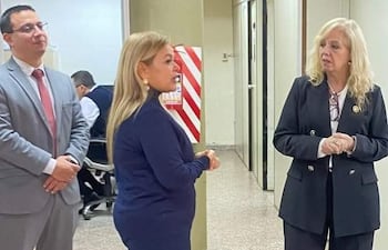 Mujeres en trajes formales discuten en ambiente de oficina. Una lleva abrigo azul oscuro y la otra traje negro.