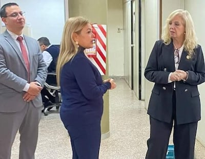 Mujeres en trajes formales discuten en ambiente de oficina. Una lleva abrigo azul oscuro y la otra traje negro.