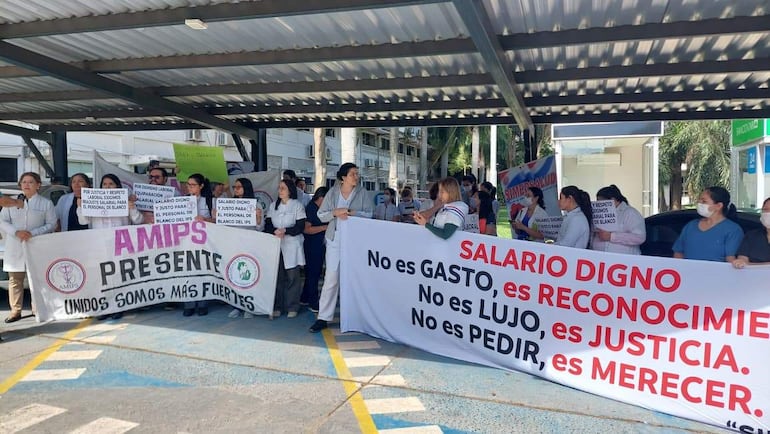 Grupo de médicos en uniformes sosteniendo pancartas que exigen 'salario digno' y 'reconocimiento' en un ambiente al aire libre.