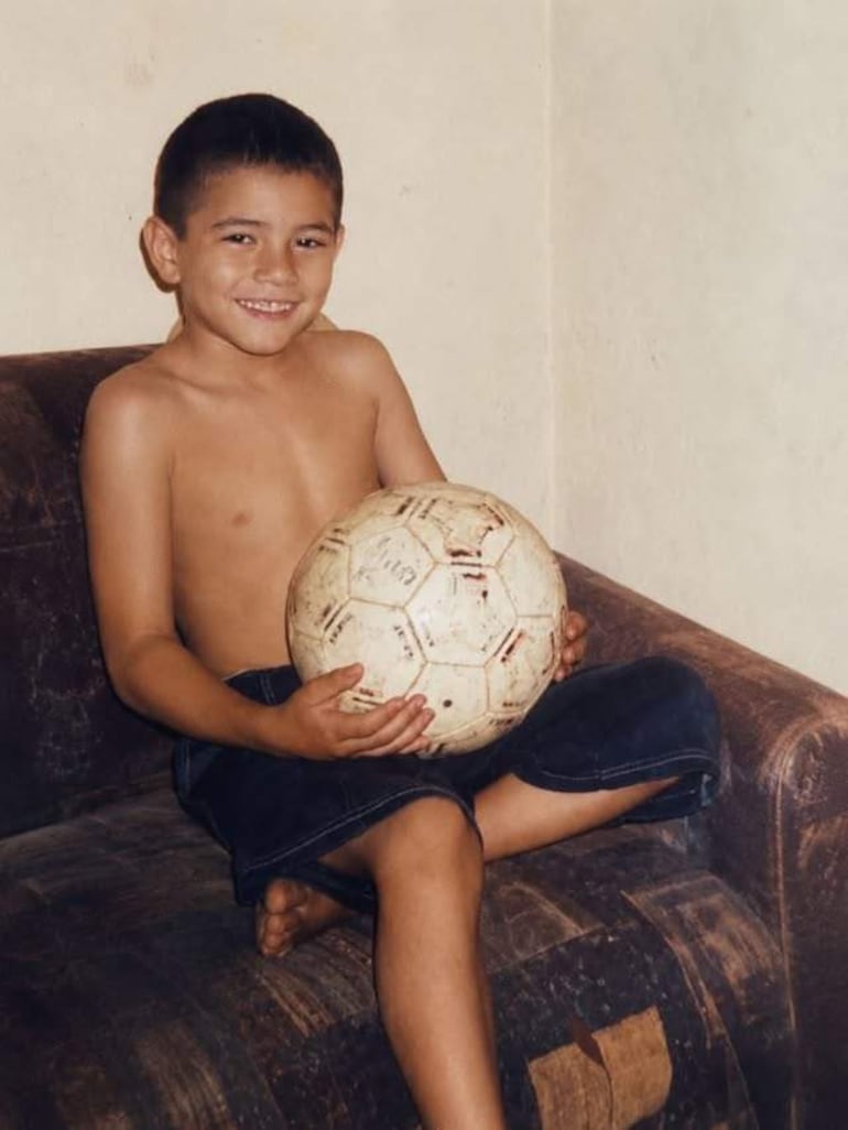 Niño de 6 a 8 años, sin camiseta y en pantalones cortos, sonríe sentado en un sofá con un balón de fútbol.
