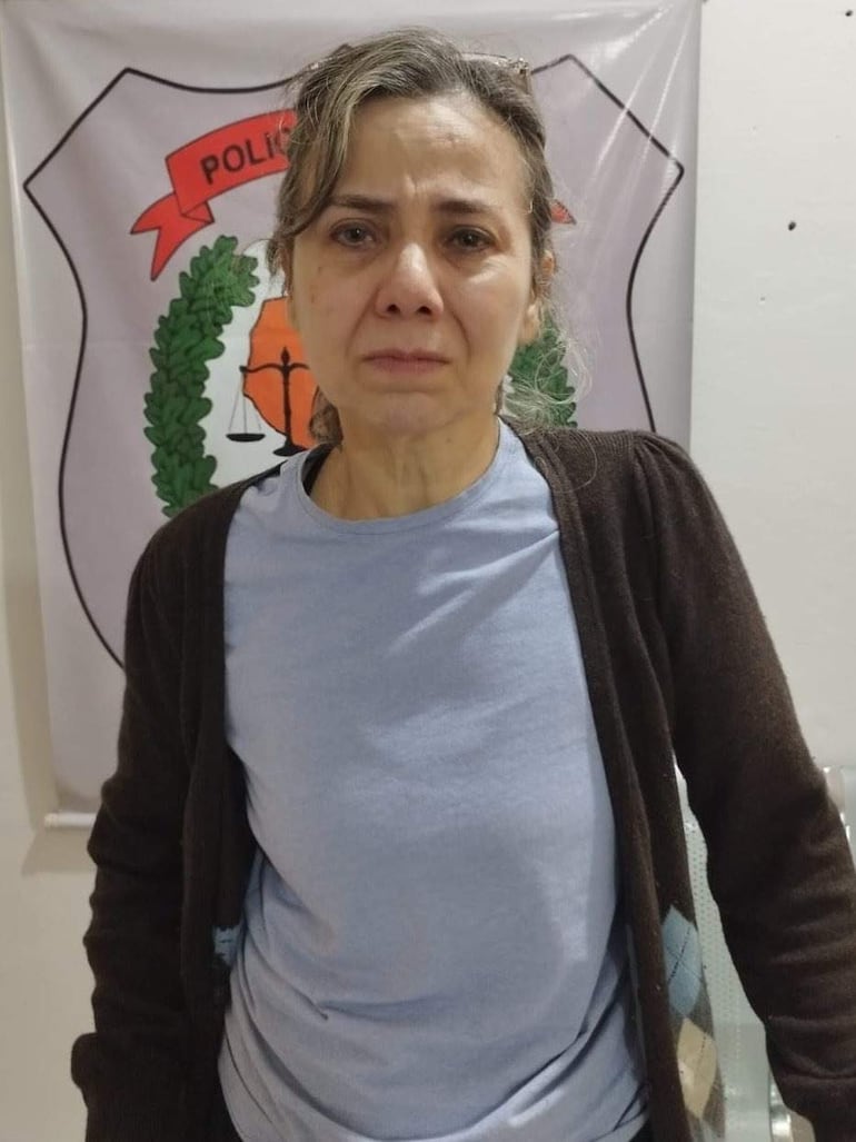 Mujer con lágrimas en los ojos, cabello suelto, vestida con camiseta clara y abrigo oscuro, frente al escudo de la Policía.