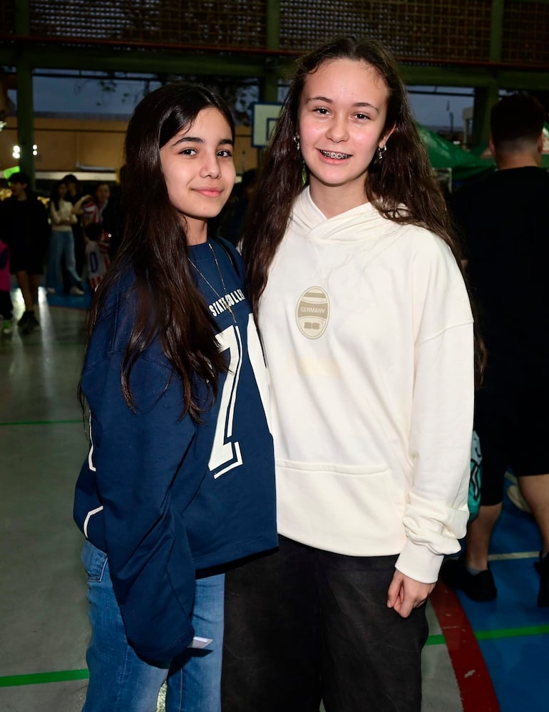 Dos chicas sonríen, una con suéter azul oscuro y otra con sudadera blanca, en un ambiente de gimnasio con iluminación artificial.