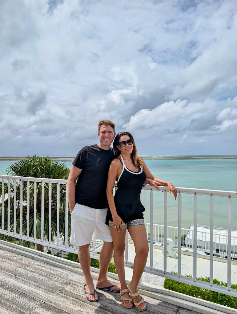 Pareja sonriente en balcón: hombre con camiseta negra y pantalones cortos blancos, mujer con atuendo negro, fondo de agua y cielo parcialmente nublado.