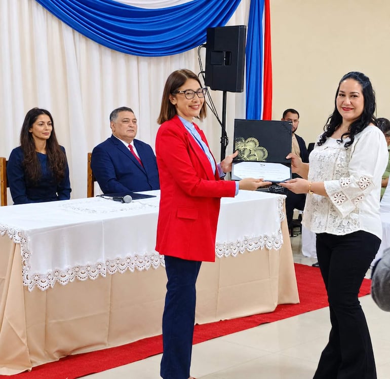 Susana Verza en blazer rojo entrega un documento con una mujer en blusa blanca. Ambiente decorado con banderas de Paraguay.