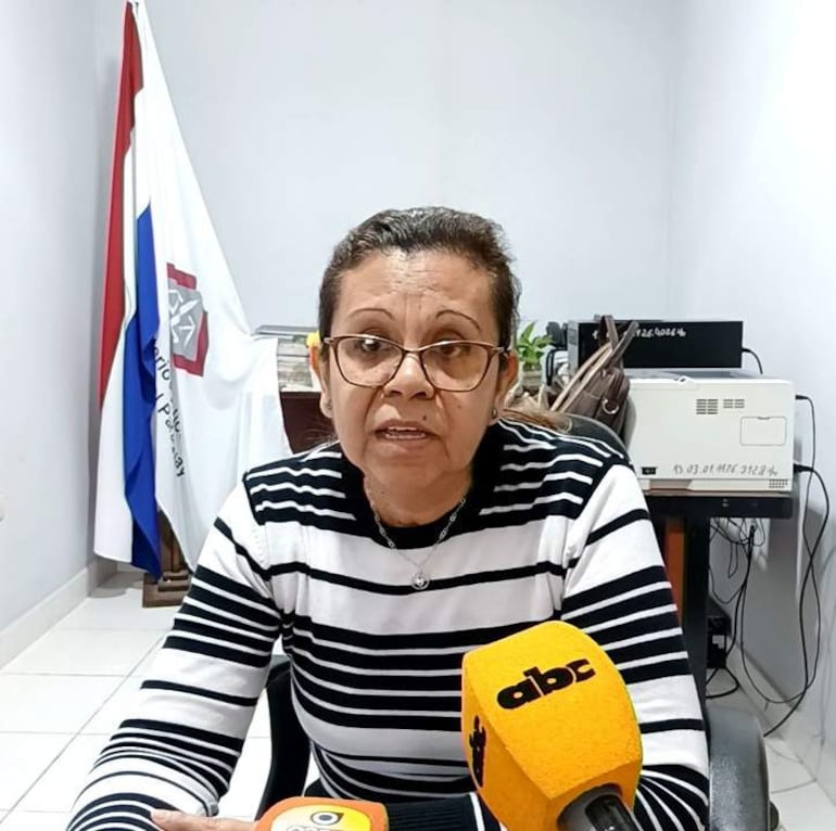 Mirtha Valle en oficina con cabello castaño claro y gafas, camiseta a rayas, rodeada de objetos de oficina y un micrófono.