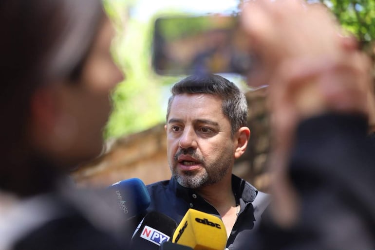 Pedro Alliana en camisa negra habla ante micrófonos; una mujer sostiene un micrófono escuchando atentamente en entorno verde.