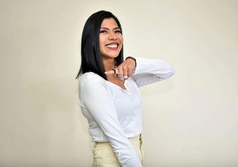 Nata Calpurnia sonríe vestida con blusa blanca y pantalones claros, apuntando hacia la cámara en un entorno interior.